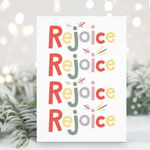 Load image into Gallery viewer, A Christmas card standing up with with pine leaves in the background with a touch of snow. The card has a white background and features the word &quot;rejoice&quot; repeated four times. The letters of the word are in different colors of muted red, yellow, green, purple and pink. 