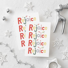 Load image into Gallery viewer, Two Christmas cards laying on a white background with white and silver Christmas decorations on the table. The card has a white background and features the word &quot;rejoice&quot; repeated four times. The letters of the word are in different colors of muted red, yellow, green, purple and pink.
