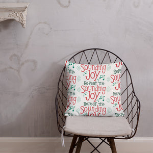 The pillow is leaning on a metal chair with a cushion. The white pillow has a repeat pattern with the phrase "Repeat the sounding joy" in red and green and musical notes around the words. 