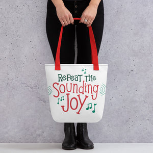 Someone holding a tote bag with red handles and a white fabric bag. The tote bag features the words "Repeat the Sounding Joy" in red and green with musical notes around the words.