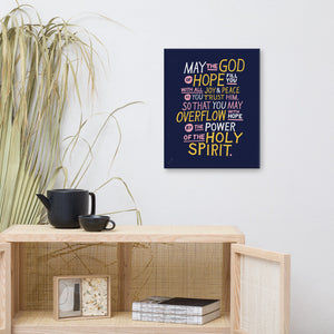 A canvas shown on a wall above a shelf. The canvas is purple and features hand drawn text in white, pink and yellow reading "May the God of hope fill you with all joy and peace as you trust him, so that you may overflow with hope by the power of the holy spirit." 