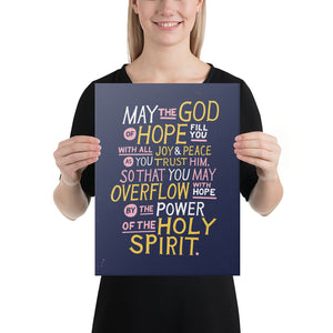 A woman holding a canvas in her hands. The canvas is purple and features hand drawn text in white, pink and yellow reading "May the God of hope fill you with all joy and peace as you trust him, so that you may overflow with hope by the power of the holy spirit." 