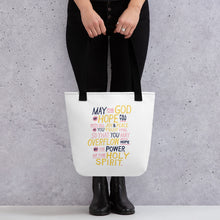 Load image into Gallery viewer, Someone holding a tote bag with black handles and a white fabric bag. The artwork features lettering in purple, pink and yellow reading &quot;May the God of hope fill you with all joy and peace as you trust him, so that you may overflow with hope by the power of the holy spirit.&quot; 
