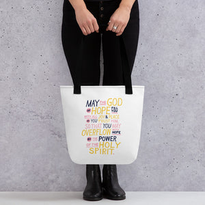 Someone holding a tote bag with black handles and a white fabric bag. The artwork features lettering in purple, pink and yellow reading "May the God of hope fill you with all joy and peace as you trust him, so that you may overflow with hope by the power of the holy spirit." 