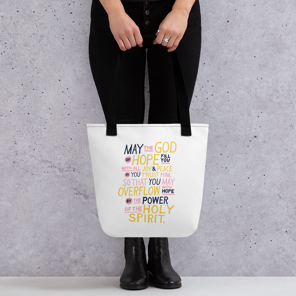 Someone holding a tote bag with black handles and a white fabric bag. The artwork features lettering in purple, pink and yellow reading 