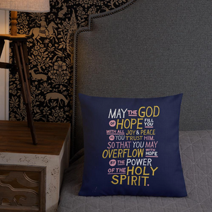 A pillow leaning on a grey headboard with a plant in the background. The purple pillow features hand drawn lettering of the Bible verse "May the God of hope fill you with all joy and peace as you trust him, so that you may overflow with hope by the power of the holy spirit." The lettering in white, pink and yellow. 
