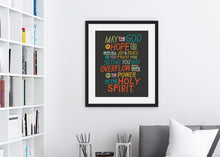 Load image into Gallery viewer, Artwork in a black frame with the with a white matte shown hanging on a wall above a sofa. The artwork is on a black background with colorful letters reading May the God of hope fill you with all joy and peace as you trust him, so that you may overflow with hope by the power of the holy spirit.&quot;
