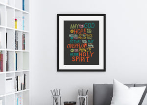 Artwork in a black frame with the with a white matte shown hanging on a wall above a sofa. The artwork is on a black background with colorful letters reading May the God of hope fill you with all joy and peace as you trust him, so that you may overflow with hope by the power of the holy spirit."