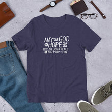 Load image into Gallery viewer, A short sleeved T-shirt laying flat with objects around it. The tee is a heather midnight blue color and features hand drawn lettering in white with the words &quot;May the God of hope fill you with all joy and peace as you trust him.&quot;