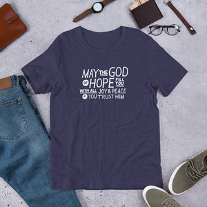 A short sleeved T-shirt laying flat with objects around it. The tee is a heather midnight blue color and features hand drawn lettering in white with the words "May the God of hope fill you with all joy and peace as you trust him."