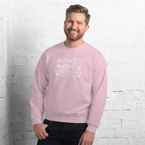 A man wearing a light pink sweatshirt featuring hand drawn lettering in white with the words "May the God of hope fill you with all joy and peace as you trust him."