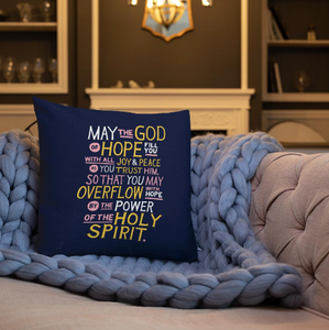 A white pillow on a sofa with a blue knitted blanket. The purple pillow features hand drawn lettering of the Bible verse "May the God of hope fill you with all joy and peace as you trust him, so that you may overflow with hope by the power of the holy spirit." The lettering in white, pink and yellow. 
