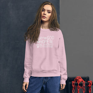 A woman wearing a light pink sweatshirt featuring hand drawn lettering in white with the words "May the God of hope fill you with all joy and peace as you trust him."