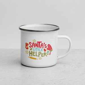 White enamel mug featured on a white table top and white background. The design features illustrated words and baking illustrations in the colors red, light blue and green. The words read "Santa's Little Helper."