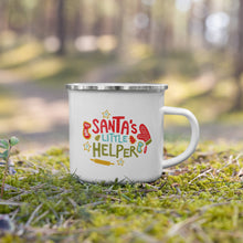 Load image into Gallery viewer, A white enamel mug with a silver enamel rim sitting in the grass. The illustrated design says &quot;Santa&#39;s Little Helper&quot; with baking illustrations around it. The design is in red, light blue and green. 