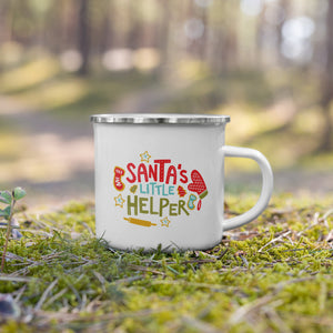 A white enamel mug with a silver enamel rim sitting in the grass. The illustrated design says "Santa's Little Helper" with baking illustrations around it. The design is in red, light blue and green. 