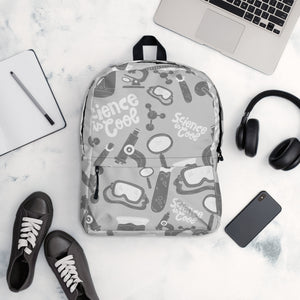 A backpack is placed on a table with a laptop, notebook, shoes, headphones and a mobile phone. The backpack is a light gray with a pattern of illustrations in darker gray and white. The pattern of illustrations features test tubes, microscopes, magnifying glasses, protective science goggles, atom models and the words "Science is cool."