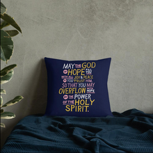Load image into Gallery viewer, A pillow leaning on a wall on a bed. The purple pillow features hand drawn lettering of the Bible verse &quot;May the God of hope fill you with all joy and peace as you trust him, so that you may overflow with hope by the power of the holy spirit.&quot; The lettering in white, pink and yellow.