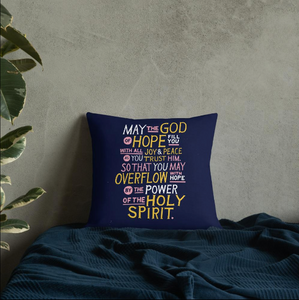 A pillow leaning on a wall on a bed. The purple pillow features hand drawn lettering of the Bible verse "May the God of hope fill you with all joy and peace as you trust him, so that you may overflow with hope by the power of the holy spirit." The lettering in white, pink and yellow.
