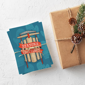 A stack of Christmas cards with brown string wrapped around them. A brown craft paper gift is off to the side. The card has a blue background with lighter blue winter mittens in a pattern. On top of the mittens is an illustrated vintage sled with red ribbon and the words "season's greetings" in white.