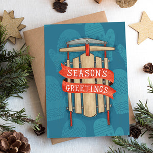 A photo of a Christmas card on top of a brown paper wrapped gift with Christmas decor around it. The card has a blue background with lighter blue winter mittens in a pattern. On top of the mittens is an illustrated vintage sled with red ribbon and the words "season's greetings" in white. 