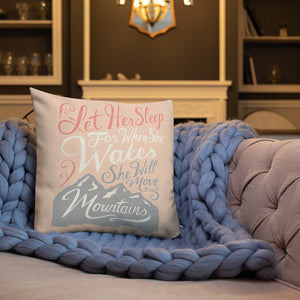 A pink cushion sits on a living room sofa with a chunky knitted purple blanket. The cushion reads 'Let her sleep for when she wakes she will move mountains' in a pink, white, and light grey lettering design, with a grey mountain illustration at the bottom.