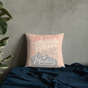 A pink cushion leans against a grey plaster wall on a navy blue bed. The cushion reads 'Let her sleep for when she wakes she will move mountains' in a pink, white, and light grey lettering design, with a grey mountain illustration at the bottom.