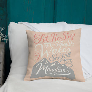 A pink cushion sits on a white sofa. The cushion reads 'Let her sleep for when she wakes she will move mountains' in a pink, white, and light grey lettering design, with a grey mountain illustration at the bottom.