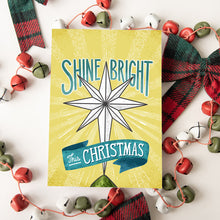 Load image into Gallery viewer, A Christmas card featured on top of some red and white Christmas decorations. The Christmas card has a yellow background with the words &#39;shine bright this Christmas&#39; in blue and white. There&#39;s an illustrated vintage star Christmas tree topper featured in between the words.