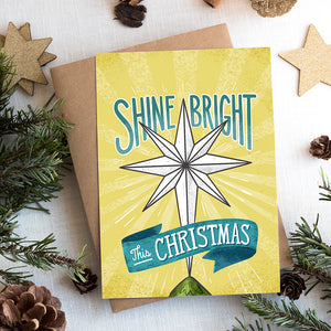 A photo of a Christmas card on top of a brown paper wrapped gift with Christmas decor around it. The Christmas card has a yellow background with the words 'shine bright this Christmas' in blue and white. There's an illustrated vintage star Christmas tree topper featured in between the words.