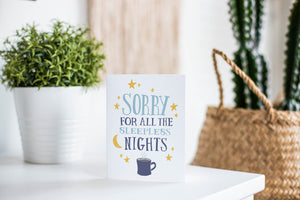 A greeting card is featured on a white tabletop with a white planter in the background with a green plant. There’s a woven basket in the background with a cactus inside. The card features the words “Sorry for all the sleepless nights.”