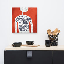 Load image into Gallery viewer, A red wall art canvas hangs on a white kitchen wall. The canvas design features the outline of a person in white, filled with light grey doodles. The words ‘There’s something in you the world needs&#39; are lettered in black across the person’s chest. Below the canvas is a black kitchen unit with a wooden worktop, holding black cups and wooden utensils.