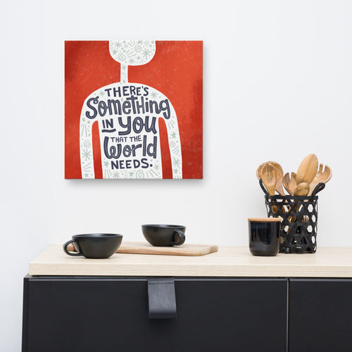 A red wall art canvas hangs on a white kitchen wall. The canvas design features the outline of a person in white, filled with light grey doodles. The words ‘There’s something in you the world needs' are lettered in black across the person’s chest. Below the canvas is a black kitchen unit with a wooden worktop, holding black cups and wooden utensils.