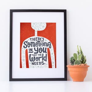 A red print in a black frame sits against a white wall. The print features the outline of a person in white, filled with light grey doodles. The words ‘There’s something in you the world needs' are lettered in black across the person’s chest. Next to the print is a small cactus in a terracotta pot..
