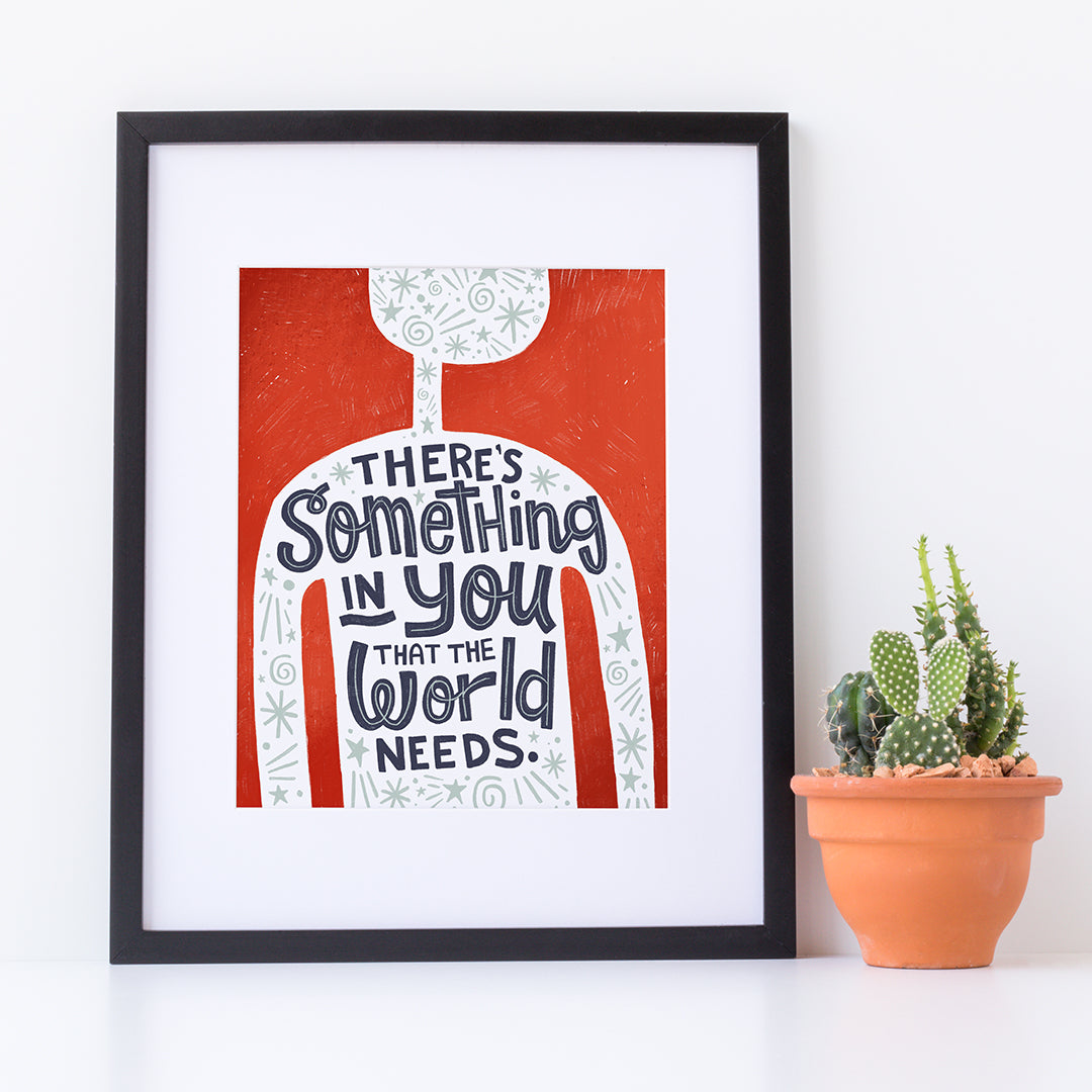 A red print in a black frame sits against a white wall. The print features the outline of a person in white, filled with light grey doodles. The words ‘There’s something in you the world needs' are lettered in black across the person’s chest. Next to the print is a small cactus in a terracotta pot..