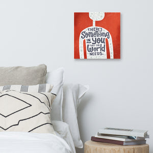A red wall art canvas hangs on a white bedroom wall. The canvas design features the outline of a person in white, filled with light grey doodles. The words ‘There’s something in you the world needs' are lettered in black across the person’s chest. Below the canvas is a white bed with lots of pillows, and a wooden sidetable holding a stack of books.
