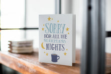 Load image into Gallery viewer, A card on a wood tabletop with an object in the background that is out of focus. The card features the words “Sorry for all the sleepless nights.”