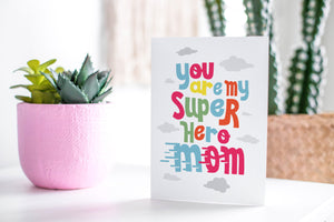 A greeting card featured standing up on a white tabletop with a pink plant pot in the background and some succulents in the pot. There’s a woven basket in the background with a cactus inside. The card features the words “You are my super hero mom.”