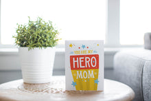 Load image into Gallery viewer, A photo of a card featured on a tabletop next to a white planter filled with a green plant. ​​The card features the words “You are my hero mom.”