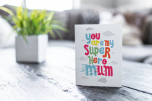 Load image into Gallery viewer, A greeting card featured on a black, wood coffee table. There’s a white planter in the background with a green plant. There’s also a gray sofa in the background with a white pillow. The card features the words “You are my super hero mum” with clouds in the background. 