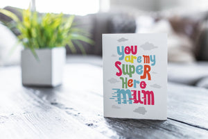 A greeting card featured on a black, wood coffee table. There’s a white planter in the background with a green plant. There’s also a gray sofa in the background with a white pillow. The card features the words “You are my super hero mum” with clouds in the background. 