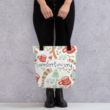 Load image into Gallery viewer, Someone holding a tote bag with black handles and a white fabric bag. The artwork features an illustrated Christmas pattern with hot cocoa mugs, scarves and hats, mittens and holy leaves. The colors featured on the white tote bag are light blue, light green and red with the words in black. 