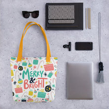 Load image into Gallery viewer, A tote bag lying on a surface with a laptop and office items next to it. The patterned tote bag has a yellow handle and the tote is white. The pattern includes the words Merry &amp; Bright in the middle with an illustrated pattern around the words with presents, ornaments, starts and candy canes. The colors of the words and patterns are in pink, light blue, yellow and black. 