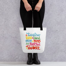 Load image into Gallery viewer, A woman holding a tote bag with black handles and the tote bag is a white/off white color. The design of the quote “A teacher takes a hand, opens a mind, and touches a heart” is featured on the bag. The “a” in the word “heart” is a heart shape and the words are blue, red, yellow and green. 