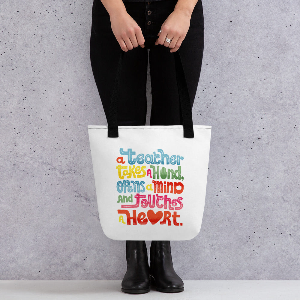 A woman holding a tote bag with black handles and the tote bag is a white/off white color. The design of the quote “A teacher takes a hand, opens a mind, and touches a heart” is featured on the bag. The “a” in the word “heart” is a heart shape and the words are blue, red, yellow and green. 