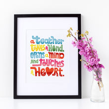 Load image into Gallery viewer, A framed print with the phrase “A teacher takes a hand, opens a mind and touches a heart.” The words are from hand lettered art and are in colorful colors. The word heart features a heart shape in place of the “a.”