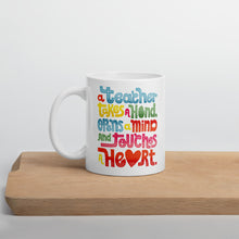 Load image into Gallery viewer, A white mug sitting on a piece of wood. The quote on the mug says “A teacher takes a hand, opens a mind, and touches a heart.” The “a” in the word “heart” is a heart shape and the words are blue, red, yellow and green. 