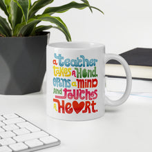 Load image into Gallery viewer, A mug featured on a desk with a plant and a keyboard. The white mug features this phrase in the colors blue, red, yellow and green. The words say “A teacher takes a hand, opens a mind, and touches a heart.”