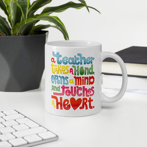 A mug featured on a desk with a plant and a keyboard. The white mug features this phrase in the colors blue, red, yellow and green. The words say “A teacher takes a hand, opens a mind, and touches a heart.”