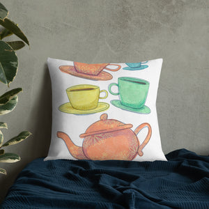 A pillow leaning on a wall on a bed with dark blue bedding. A pillow on a chair against a grey wall. The white pillow features the artwork on a white background with four teacups on saucers and one large teapot. The teacups are in muted colors of orange, blue, yellow and green and the teapot is a muted orange. On the teacups, saucers and teapot there is a light flower detail pattern.